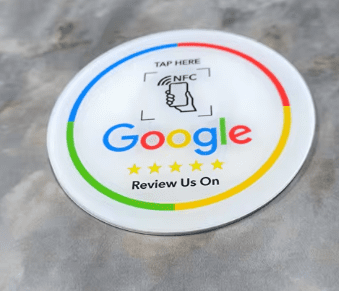 A round white sign featuring Google's logo in the center with the text "TAP HERE" at the top and a contactless NFC symbol. Below the logo, the text "Review Us On" and five yellow stars are displayed, encircled by blue, green, yellow, and red borders.