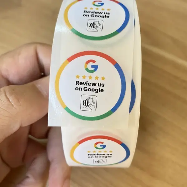 A hand holds a roll of round stickers with a Google logo and stars. The stickers read "Review us on Google" and display a tapping gesture with an NFC symbol, suggesting the use of NFC TAP CARDS for easy review access.
