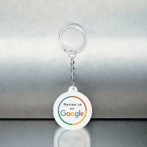 A white keychain with a round medallion hangs against a metallic background. The medallion features the text "Review us on Google," with the word "Google" in its distinctive multi-color logo font. The keychain is reflected on the shiny surface it rests on.