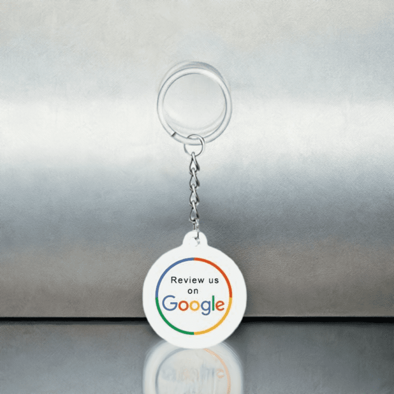 A white keychain with a round medallion hangs against a metallic background. The medallion features the text "Review us on Google," with the word "Google" in its distinctive multi-color logo font. The keychain is reflected on the shiny surface it rests on.