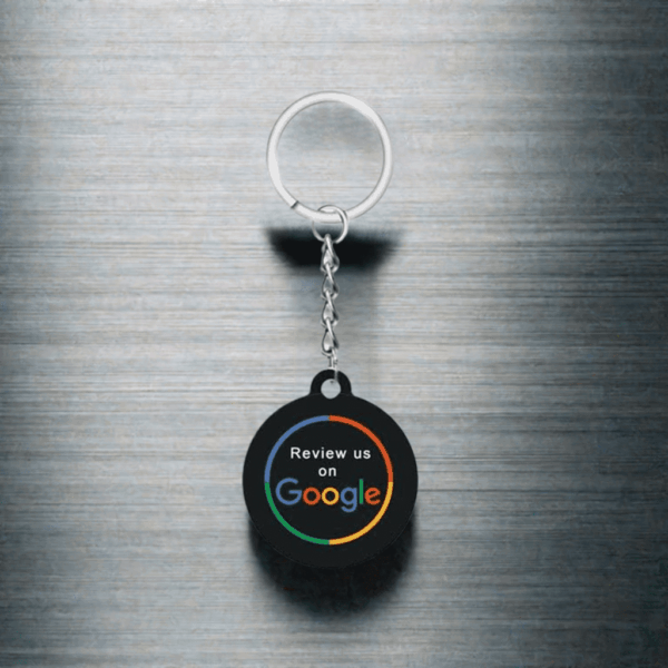 A round NFC keychain with a metal ring and chain is displayed against a brushed metal background. The keychain features the text "Review us on Google," with the Google logo colors in the word "Google.
