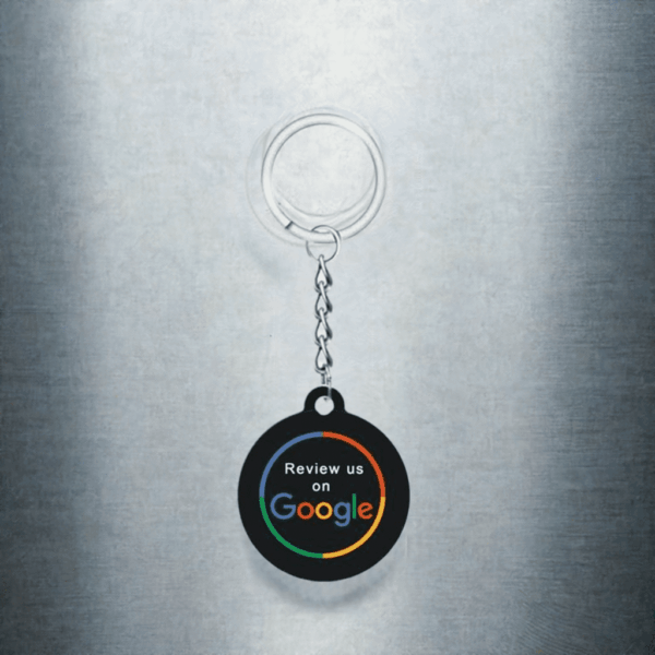 A keychain hangs against a plain, metallic background. The keychain has a circular black tag with the phrase "Review us on Google" printed in white text, featuring the colorful Google logo. This NFC keychain for Google reviews comes with a metallic silver keyring and an attached short chain.