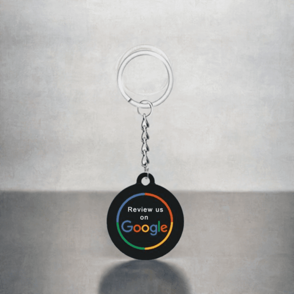 A keychain with a circular black tag hangs against a gray background. The tag reads "Review us on Google" in colorful letters, hinting at its functionality. This NFC keychain for Google reviews is attached to a metallic keyring through a small chain.