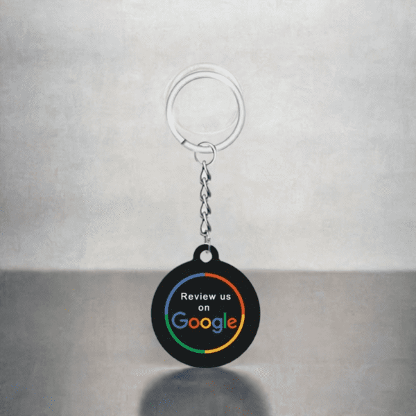 A circular black keychain with text reading "Review us on Google" in white and Google in its signature colors, attached to a metal keyring and chain, is placed against a light grey background. This stylish accessory is perfect for promoting your business at any NFC station.