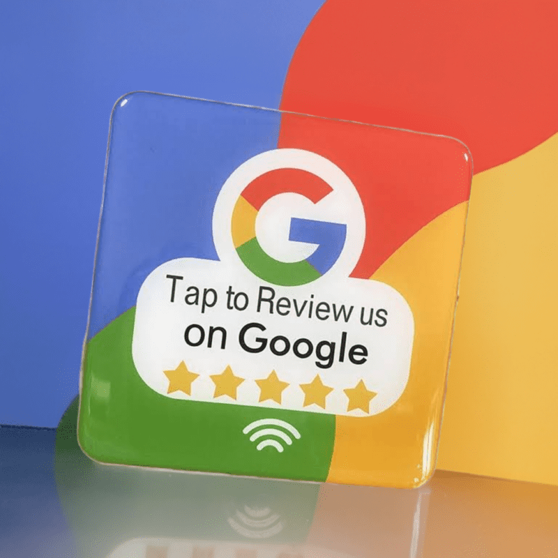 A Google review card with the iconic Google logo and the text "Tap to Review us on Google" is displayed against a vibrant background of blue, red, green, and yellow. Below the prompt are five gold stars. This card also functions as an NFC station for ease of access.