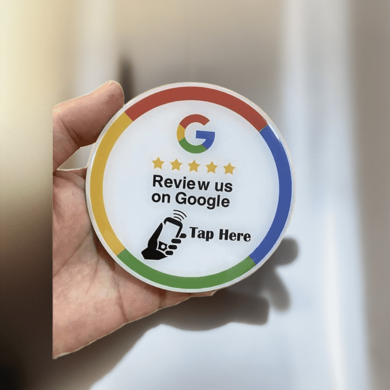 A person's hand holding a round sticker with Google's logo at the top, five stars, the text "Review us on Google," and an icon of a hand tapping a smartphone, indicating where to tap. The sticker is surrounded by a colorful border in red, yellow, green, and blue.