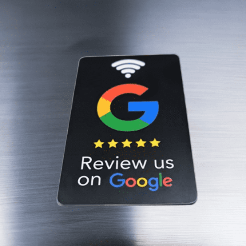 A card lays on a metallic surface, featuring a large multicolored "G" logo with five yellow stars underneath. Above the "G" is a Wi-Fi symbol. The text below reads "Review us on Google" in white.